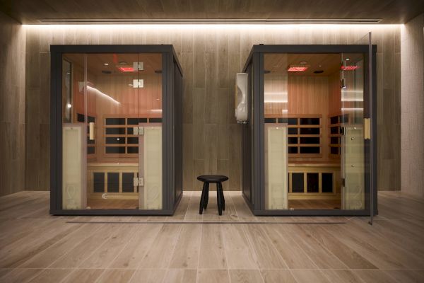 The image shows two modern glass sauna rooms with a wooden interior and a black stool placed between them on a wooden floor.