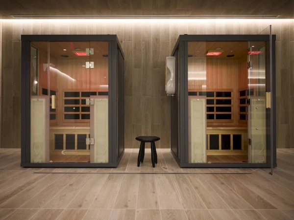 The image shows two modern glass sauna rooms with a wooden interior and a black stool placed between them on a wooden floor.