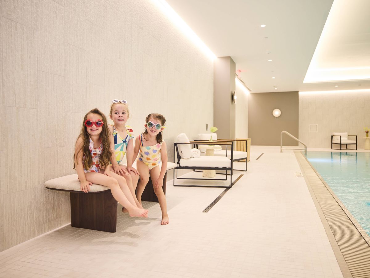 Three children in swimsuits and goggles sit on a bench by an indoor pool.