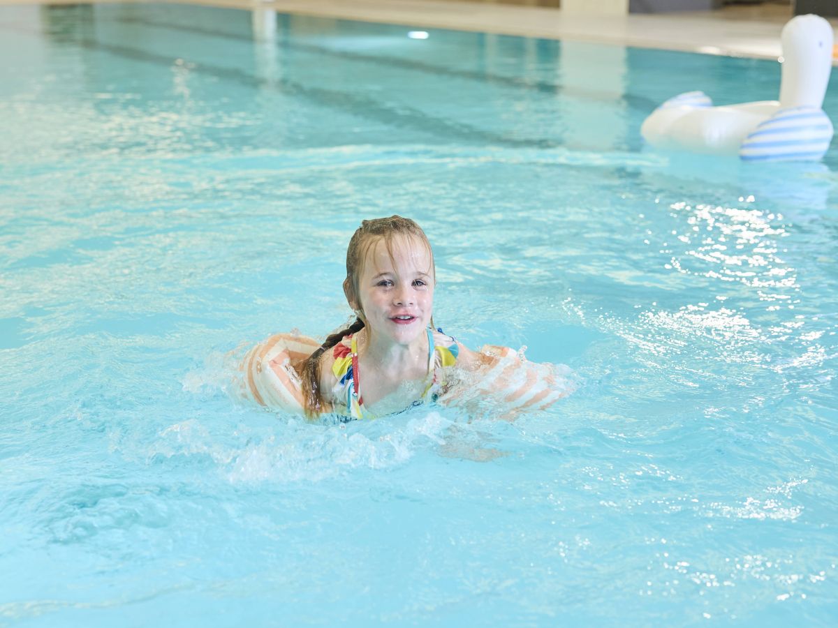 A child is swimming in a pool wearing arm floaties, with a swan pool float in the background.