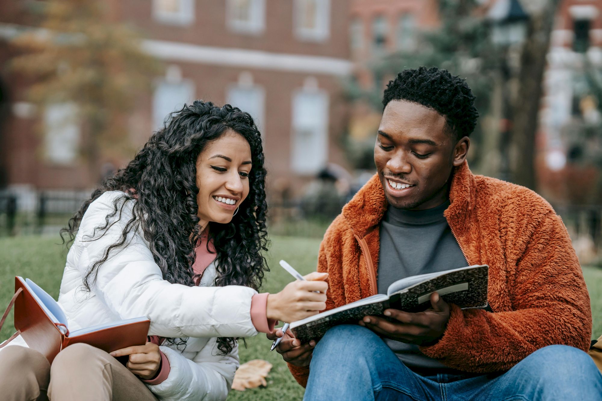 Two people are sitting on grass, studying together and smiling, with books in their hands.