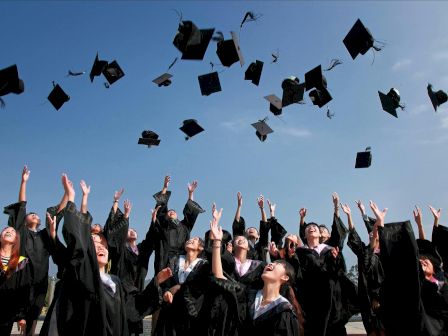 Graduates in caps and gowns celebrating by tossing their caps into the air against a clear blue sky.