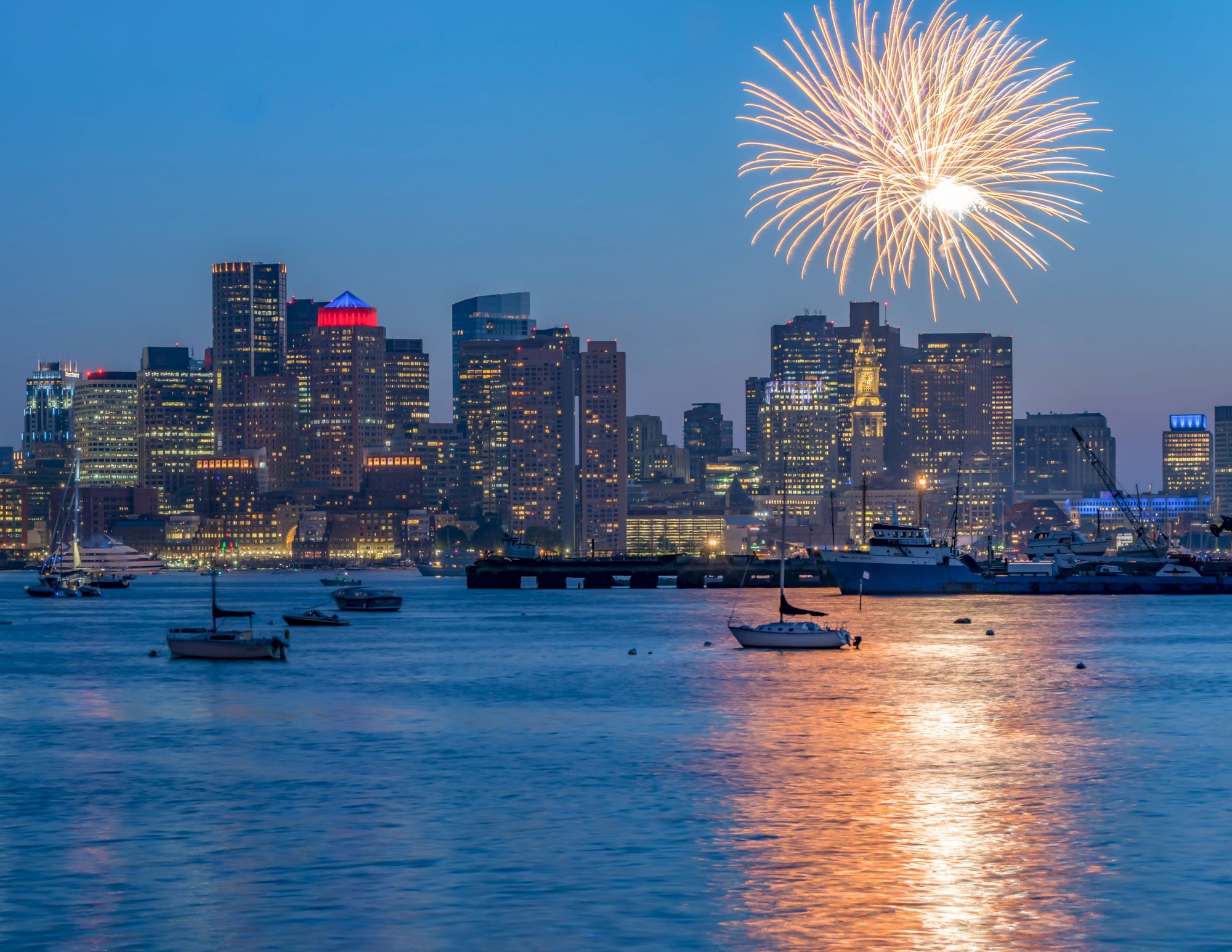A city skyline at dusk with fireworks above and boats on the water beneath, reflecting lights on the surface.