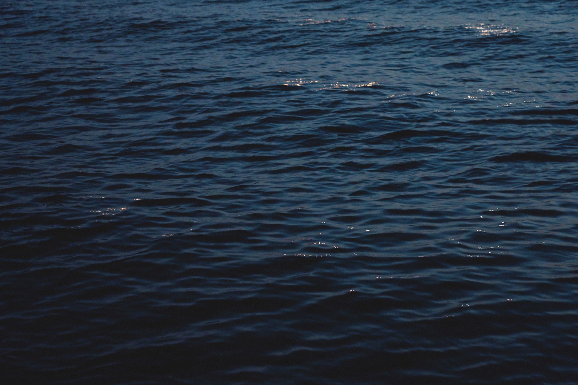 A dark ocean with rippling waves and scattered distant city lights shimmering on the water at night.