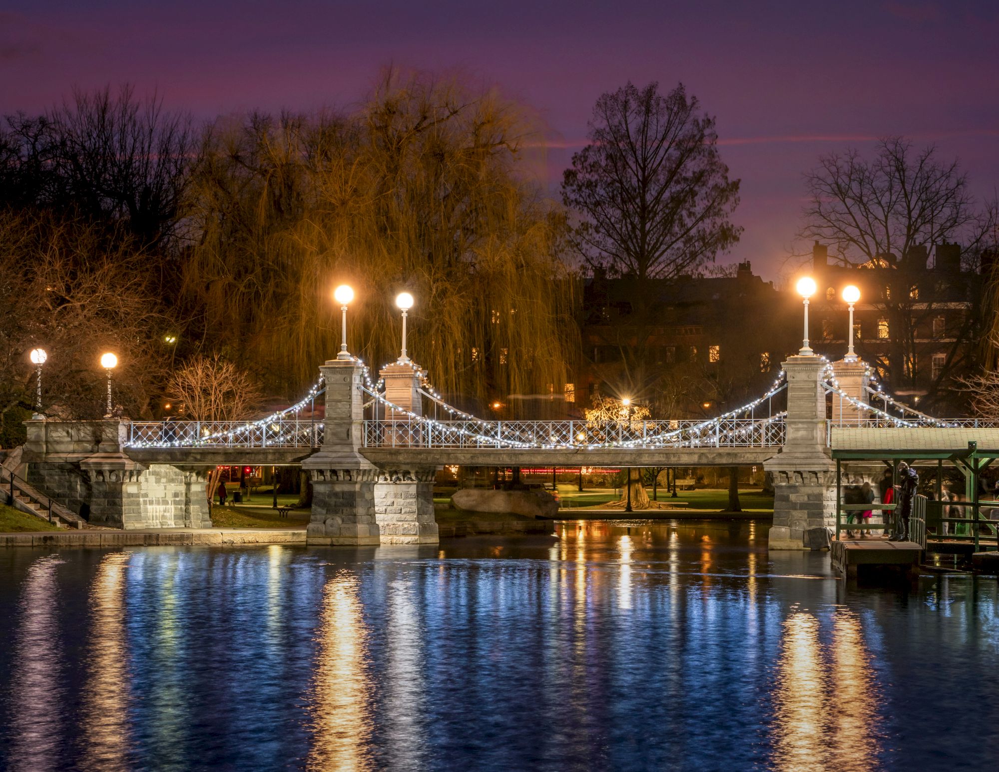 A beautifully illuminated bridge over a calm body of water at dusk, with trees and buildings in the background, reflecting warm lights.