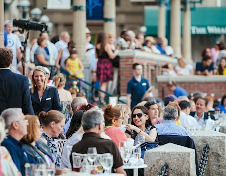 A lively outdoor gathering with people dining and socializing at tables, surrounded by a bustling crowd in a festive atmosphere.