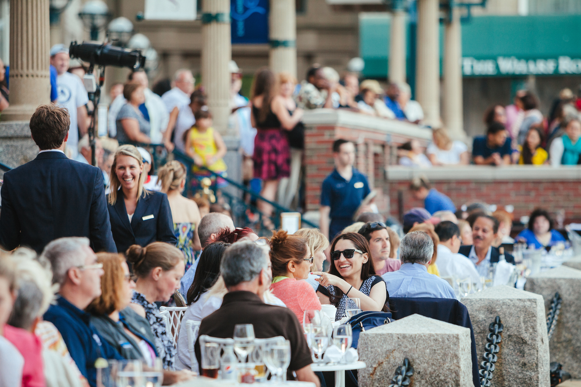 A lively outdoor gathering with people dining and socializing at tables, surrounded by a bustling crowd in a festive atmosphere.