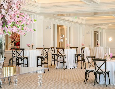 The image shows an elegant event setup with tall tables covered in white cloth, black chairs, and pink floral arrangements in a bright room.