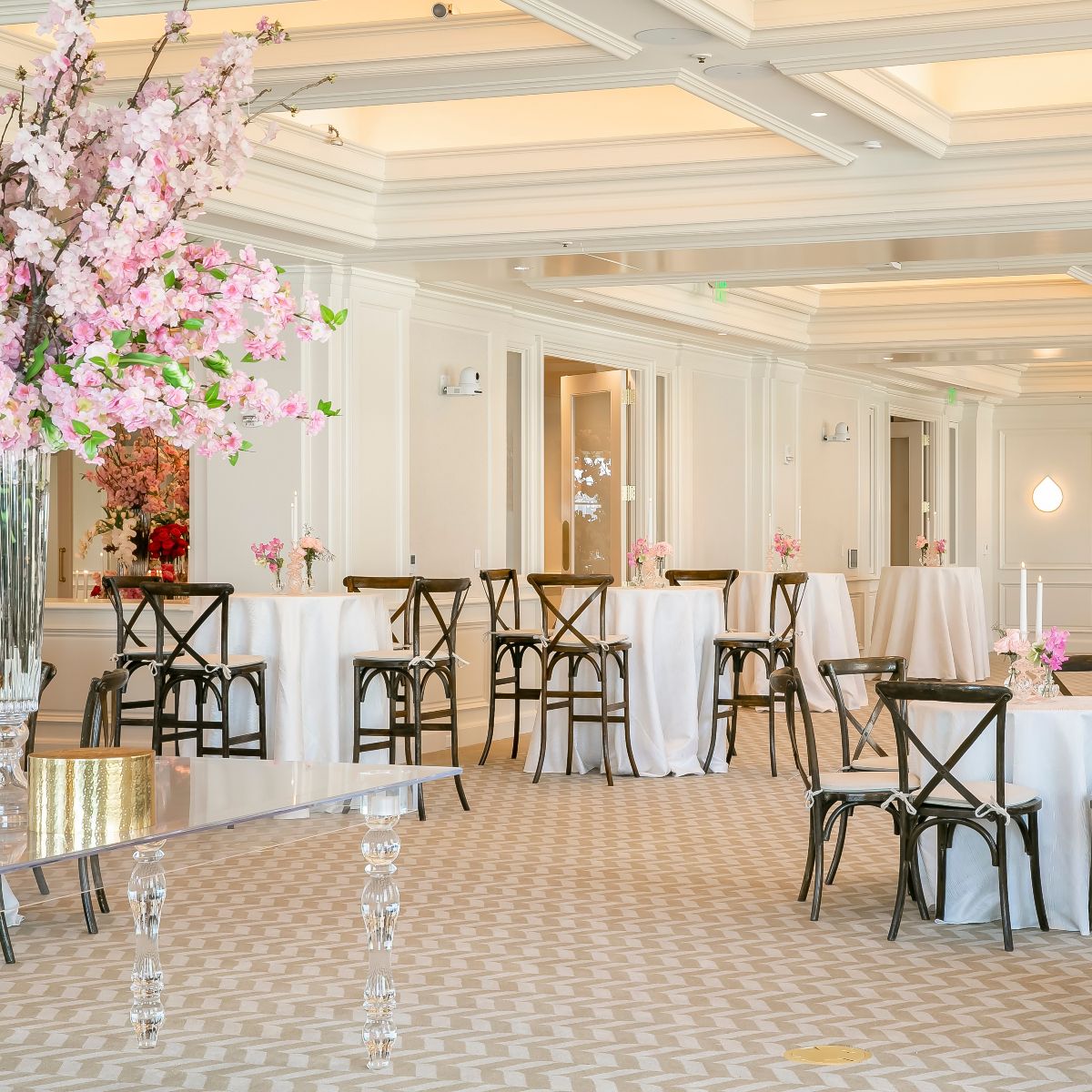 The image shows an elegant event setup with tall tables covered in white cloth, black chairs, and pink floral arrangements in a bright room.