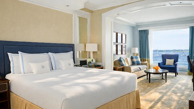 A bright hotel suite with a large bed, beige walls, and a blue headboard; sitting area with blue chair, coffee table, and sunny balcony view.