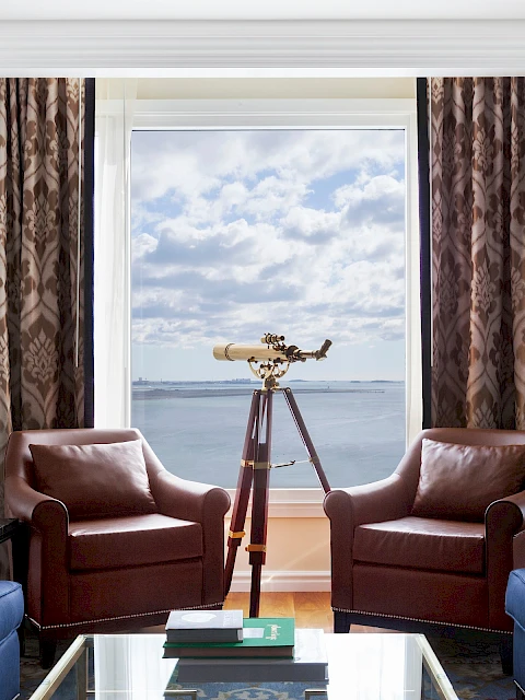 Luxurious room with a telescope by the window, blue sofas, brown chairs, and decorative lamps; sea view with cloudy sky.