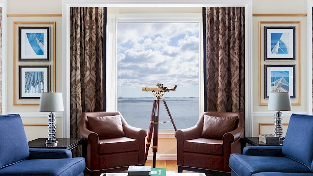 Luxurious room with a telescope by the window, blue sofas, brown chairs, and decorative lamps; sea view with cloudy sky.