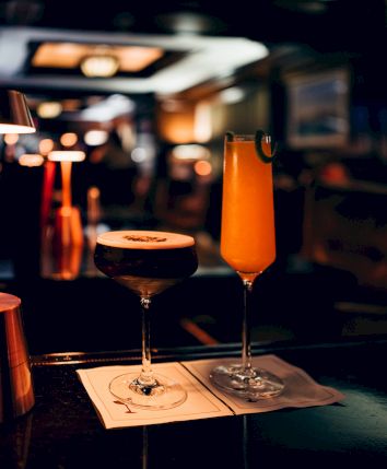 Two cocktails on a bar counter in a dimly lit setting, one dark and frothy, the other orange with a garnish.