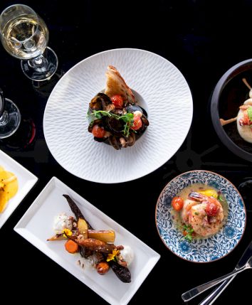 An elegant dining setup with five gourmet dishes, wine glasses, and utensils arranged on a dark table.