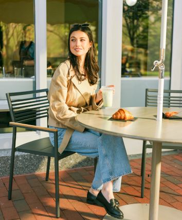 A woman sits outdoors at a café table, holding a drink, with croissants in front of her. She's wearing a beige coat and jeans.