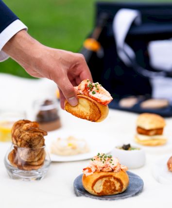A hand holding a gourmet appetizer over a table set with various small dishes and snacks, against an outdoor background.