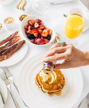 A breakfast spread with pancakes, fruit, bacon, and orange juice. A hand pours syrup on pancakes.