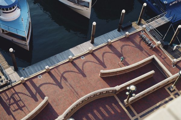 Aerial view of a waterfront promenade with boats docked, casting shadows of arches and lamps on the red-brick pathway, with one person walking.