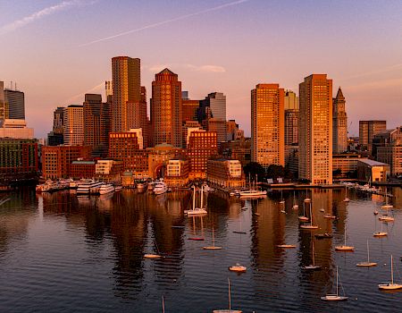 A city skyline at sunset, with skyscrapers and a harbor filled with boats reflecting the warm light on the water's surface.