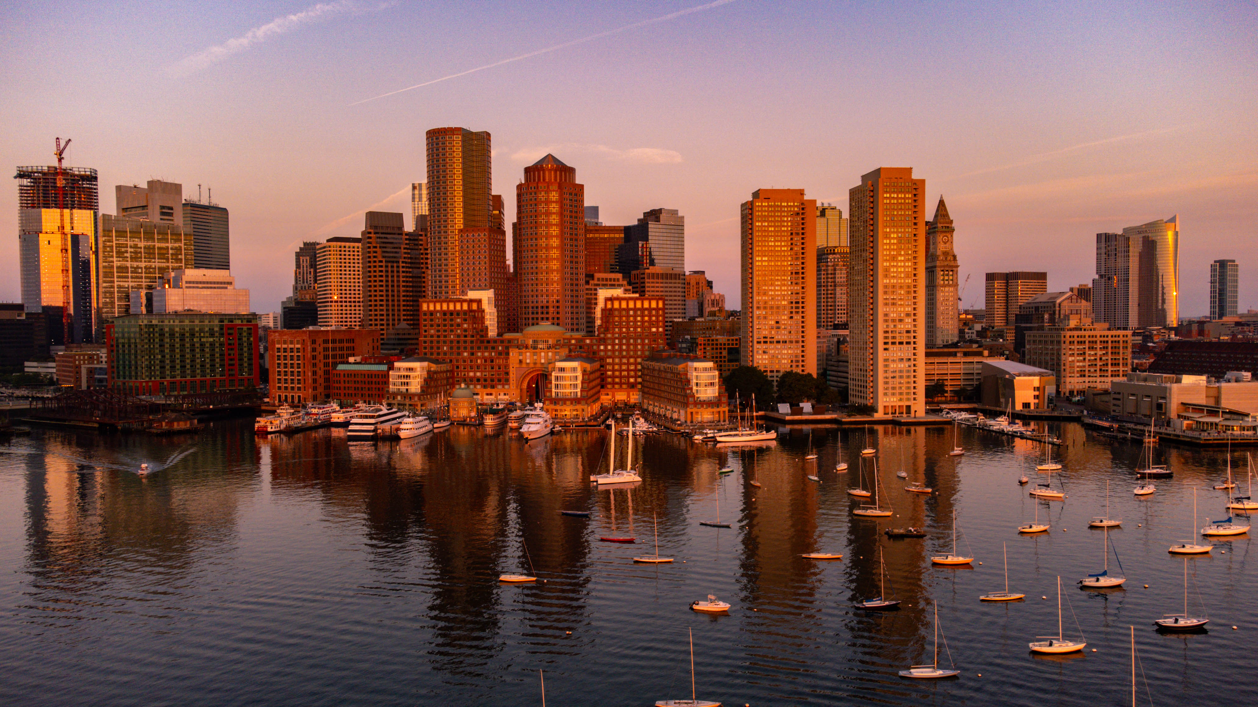 A city skyline at sunset, with skyscrapers and a harbor filled with boats reflecting the warm light on the water's surface.