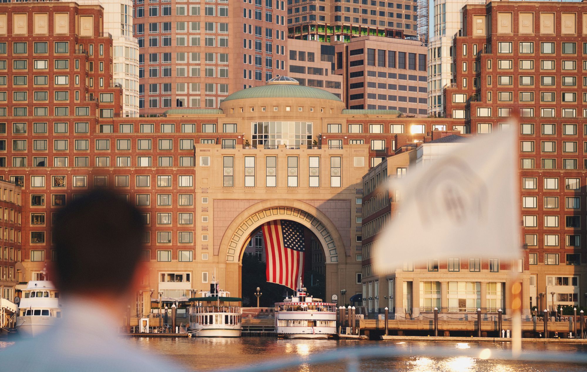 A person views a harbor with boats docked near a large building. The building displays a prominent American flag.