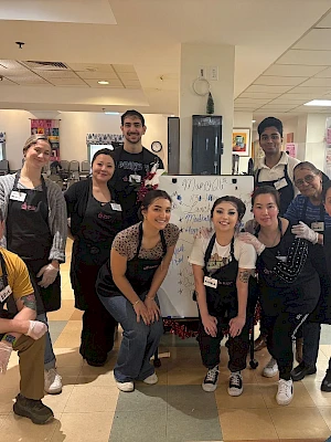 A group of people wearing aprons and gloves pose together indoors, smiling in front of a sign or menu board.