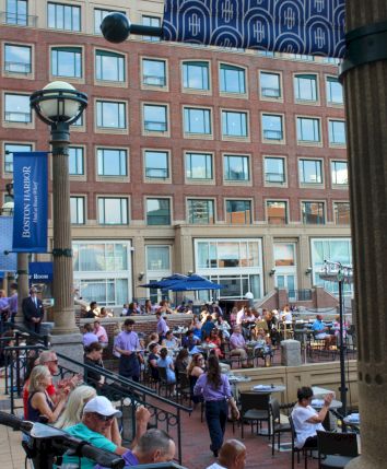 A bustling outdoor cafe scene with people seated and standing around tables, surrounded by buildings under a blue awning.