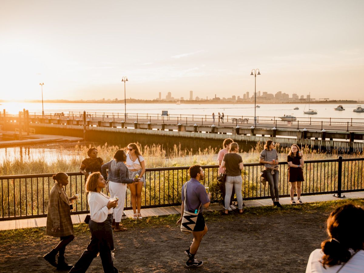 People enjoy a scenic sunset by the water with city skyline views, walking and socializing near a pier and grassy area.