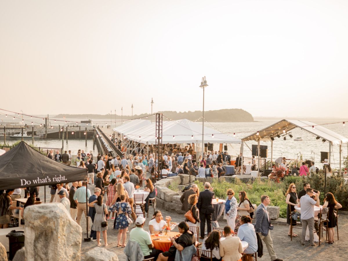 A coastal outdoor event with people mingling, seated at tables, and covered tents, set against a sea backdrop near a pier at sunset.