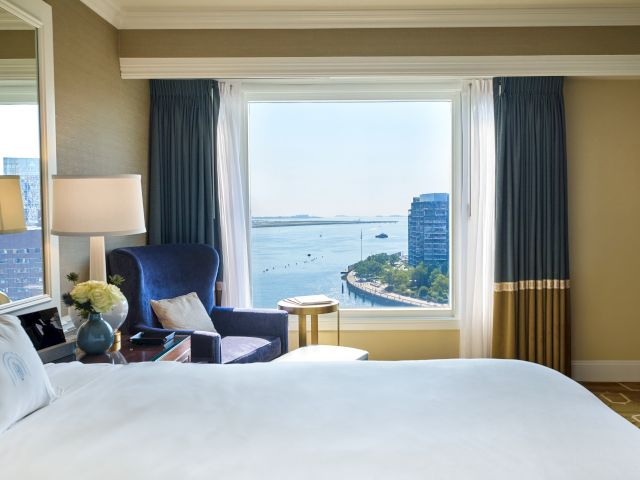 A cozy hotel room features a bed, a chair with a side table, and a window offering a stunning view of a river and buildings outside.