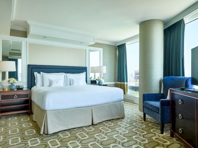 A well-lit hotel room with a large bed, side tables, lamps, a chair, and patterned carpet by a window with city views.