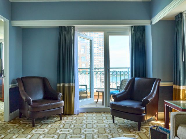 The image shows a hotel room with two leather chairs, a balcony door, and blue curtains, offering a view outside.