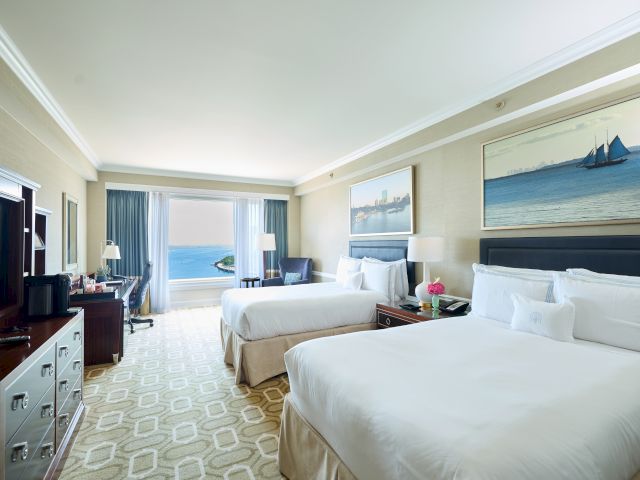 This image shows a hotel room with two beds, a TV, a dresser, and ocean view. The decor includes paintings of boats, with calming tones.