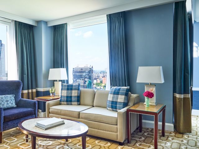 A hotel living room with a sofa, armchair, coffee table, and two lamps by the windows displaying a city view.
