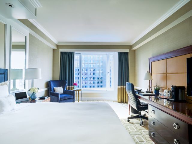A hotel room with a large bed, desk, chair, TV, and window overlooking a building is shown in the image.