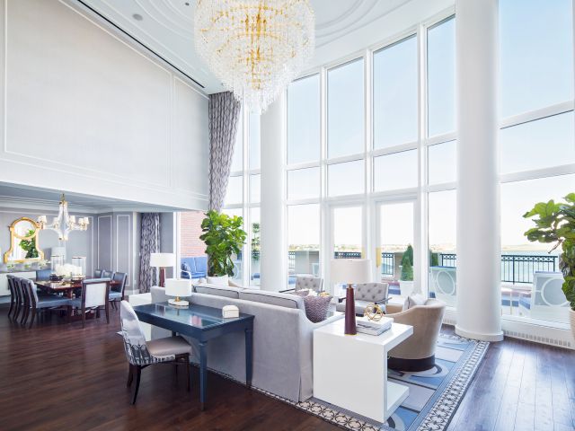 Spacious luxury living room with high ceilings, large windows, elegant furniture, a dining area, and plants, complemented by a chandelier.